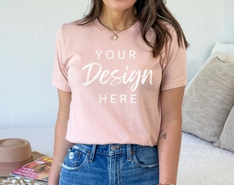Puede incluir: Camiseta rosa claro con el texto "YOUR Design HERE" en blanco. La modelo lleva vaqueros azules. Un sombrero marrón y una revista están sobre una cama con una manta a cuadros.
