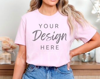 Puede incluir: Una camiseta rosa con el texto "YOUR Design HERE" impreso en la parte delantera. La camiseta la lleva puesta una persona con el pelo largo rubio y unos pantalones cortos de mezclilla azul.