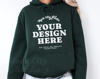 May include: A dark green hooded sweatshirt with the text "Mock City Studio" and "YOUR DESIGN HERE" in white. The text below the design reads "REAL PEOPLE - REAL PRODUCTS MOCKUP STUDIO".