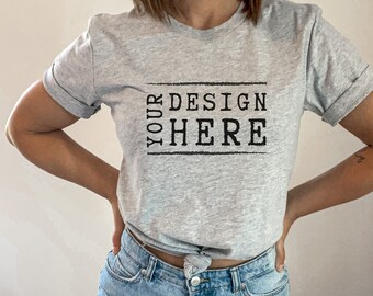 May include: Light gray t-shirt with rolled sleeves and the text "YOUR DESIGN HERE" in a black, rectangular frame. The shirt is paired with light blue, ripped jeans. The model is standing on a white, fluffy rug.