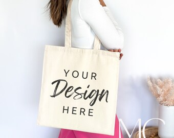May include: A person is holding a beige tote bag with the text "YOUR Design HERE" printed on it. The bag is made of canvas and has a long strap.