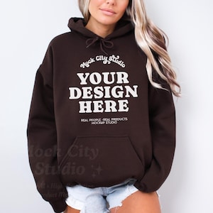Puede incluir: Una sudadera con capucha marrón con una impresión blanca que dice "Mock City Studio" y "YOUR DESIGN HERE". El texto también dice "REAL PEOPLE-REAL PRODUCTS MOCKUP STUDIO".