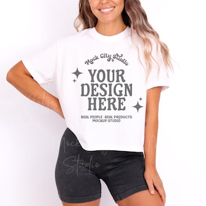 May include: White cropped t-shirt with the text "Mock City Studio" and "YOUR DESIGN HERE" in a retro font. The text is surrounded by small stars. Below the text is "REAL PEOPLE - REAL PRODUCTS MOCKUP STUDIO".