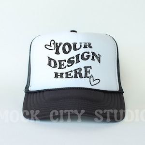 May include: Black and white trucker hat with a white mesh back. The front of the hat has the text "YOUR DESIGN HERE" in a black, distressed font with two small hearts.