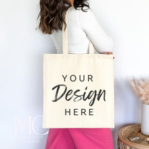 Puede incluir: Una mujer sostiene una bolsa de lona blanca con el texto "YOUR Design HERE" impreso en ella. Lleva una camisa blanca de manga larga y pantalones rosas.