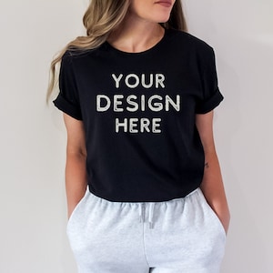 May include: A black t-shirt with the text "YOUR DESIGN HERE" printed on the front. The t-shirt is being worn by a person with long brown hair. The person is wearing grey sweatpants.