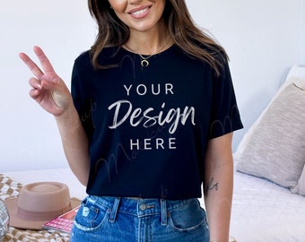 以下が含まれることがあります： 白い文字で「YOUR Design HERE」と書かれた黒いTシャツ。長い茶色の髪をした人が着ており、青いジーンズと合わせています。