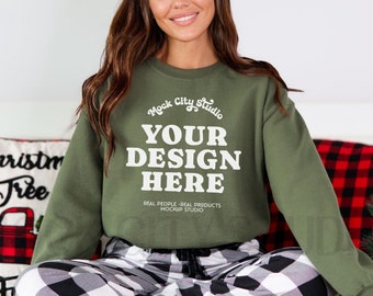 May include: A green sweatshirt with the text "Mock City Studio" and "YOUR DESIGN HERE" in white. The sweatshirt is being worn by a person sitting on a bed with a red and black plaid pillow.