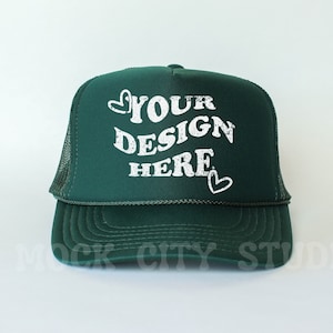 Puede incluir: Una gorra de camionero verde oscuro con una malla blanca en la parte posterior. La parte delantera de la gorra tiene texto blanco que dice "YOUR DESIGN HERE" con un corazón a cada lado del texto.