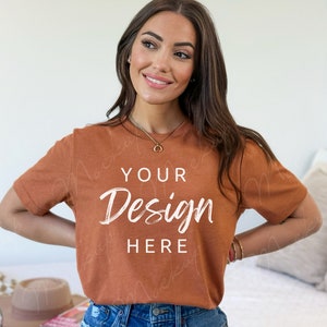 May include: A woman wearing a rust colored t-shirt with the text "YOUR Design HERE" printed on the front. She is wearing blue jeans and has long brown hair.