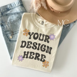 May include: A cream colored t-shirt with the text "YOUR DESIGN HERE" in black retro font. The shirt is decorated with colorful flowers and is laid out on a white surface with a pair of blue jeans and a brown hat.