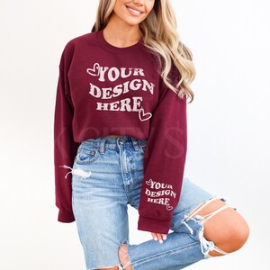 May include: A maroon cropped sweatshirt with long sleeves and the text "YOUR DESIGN HERE" in white with a heart on either side of the text. The sweatshirt has a distressed look and the text is slightly faded.