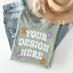Op de afbeelding: Een saliegroen T-shirt met de tekst "YOUR DESIGN HERE" in wit met een retro lettertype. Het shirt is versierd met paarse en oranje bloemen.