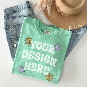 May include: A light green t-shirt with the text "YOUR DESIGN HERE" in white with yellow and purple flowers. The t-shirt is folded and laid on a white surface with blue jeans and a brown hat.