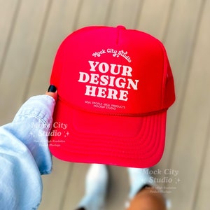 May include: A bright red trucker hat with a white mesh back. The hat has the text "Mock City Studio" and "YOUR DESIGN HERE" in white. The text "REAL PEOPLE - REAL PRODUCTS MOCKUP STUDIO" is also in white.
