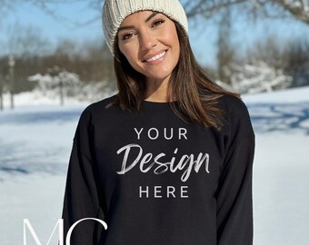 May include: A black long-sleeved sweatshirt with the text "YOUR Design HERE" in white script font. The sweatshirt is worn by a person wearing a white knit beanie with a pom-pom. The person is standing in a snowy outdoor setting.