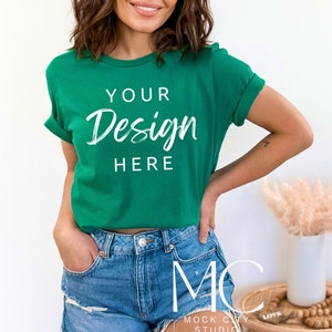 Op de afbeelding: Een vrouw draagt een groen T-shirt met de tekst "Your Design Here" in wit. Ze draagt een spijkerbroekje en heeft haar handen in haar zakken.