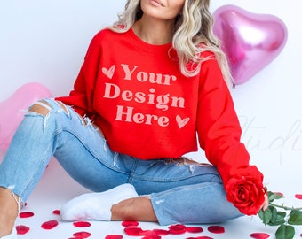 Puede incluir: Una sudadera roja con el texto "Your Design Here" en blanco, combinada con vaqueros rotos de color azul claro y zapatillas blancas. Una rosa roja y globos con forma de corazón añaden el tema de San Valentín. Pétalos de rosa rojos esparcidos en el suelo.