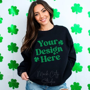 Puede incluir: Sudadera negra con el texto "Your Design Here" en verde. La sudadera está siendo modelada por una persona con cabello largo y castaño. El fondo es blanco con recortes de trébol verde.