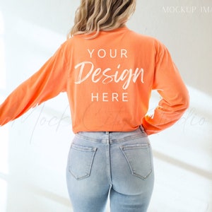 May include: A woman wearing a long-sleeved, orange t-shirt with the text "YOUR Design HERE" on the back. She is wearing blue jeans.