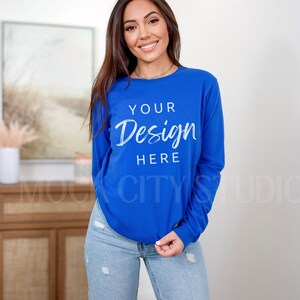 May include: A woman wearing a royal blue long-sleeved shirt with the text "YOUR Design HERE" printed on the front. She is wearing blue jeans and standing in front of a white wall.