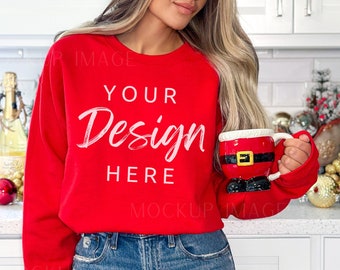 May include: A red crew neck sweatshirt with the text "YOUR Design HERE" in white. The person is holding a Santa-themed mug. The sweatshirt is paired with blue jeans. The background includes a kitchen setting with festive decorations.