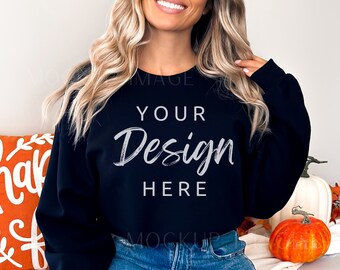 May include: A black sweatshirt with the text "YOUR Design HERE" on the front. The sweatshirt is being worn by a person with long blonde hair and blue jeans. The person is sitting on a white surface with a pumpkin and a pillow.