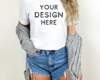 May include: A white t-shirt with the text "YOUR DESIGN HERE" printed on the front. The shirt is being worn by a person with long blonde hair and is paired with denim shorts and a gray cardigan.