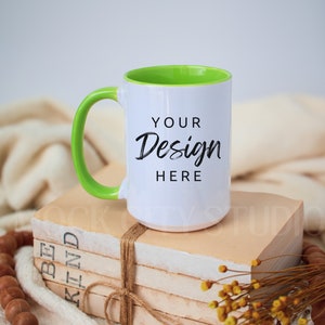 May include: White ceramic mug with a green rim and handle. The mug is sitting on a stack of books with a brown twine bow. The mug has the text "Your Design Here" written on it.