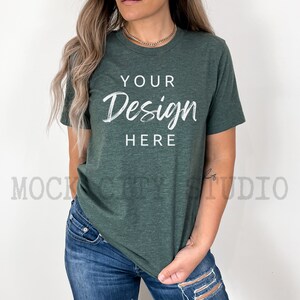 May include: A woman wearing a dark green t-shirt with the text "YOUR Design HERE" printed on the front. She is wearing blue jeans and has her hair pulled back.