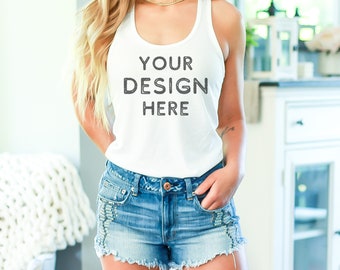 Puede incluir: Un top blanco de tirantes con el texto "YOUR DESIGN HERE" impreso en la parte delantera. El top está siendo usado por una persona con el pelo largo y rubio y se combina con unos pantalones cortos de mezclilla.