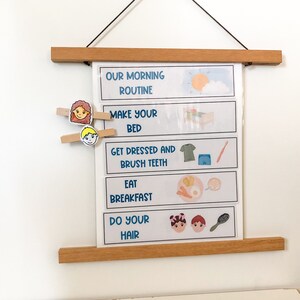 May include: A white laminated chart with a wooden frame and a blue and white illustrated morning routine for children. The chart includes the following tasks: make your bed, get dressed and brush teeth, eat breakfast, and do your hair. The chart has a clip at the top for hanging and two clothespins with cartoon faces.