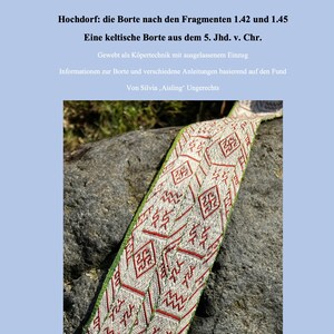 Könnte beinhalten: Nahaufnahme einer weißen, roten und grünen gewebten keltischen Borte auf einem grauen Stein. Die Borte ist eine Nachbildung eines Fundes aus dem 5. Jahrhundert v. Chr. in Hochdorf, Deutschland. Der Text "Hochdorf: die Borte nach den Fragmenten 1.42 und 1.45" ist oben im Bild sichtbar.