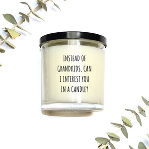 May include: A clear glass candle jar with a black lid. The candle has a white wax interior and features the text: "INSTEAD OF GRANDKIDS, CAN I INTEREST YOU IN A CANDLE?" Eucalyptus sprigs are in the background.