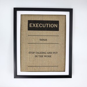 May include: Framed artwork with a black frame and white matting. The artwork features the word "EXECUTION" in a bold, black font above the word "noun" and the phrase "STOP TALKING AND PUT IN THE WORK" on a burlap background.