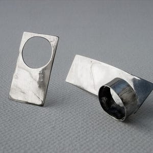 Puede incluir: Dos anillos de plata con un diseño geométrico moderno. Uno tiene forma rectangular con un círculo recortado. El otro tiene una banda rectangular con un elemento cilíndrico curvado. Los anillos se muestran sobre una superficie gris.