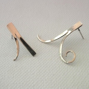 Könnte beinhalten: Ein Paar silberfarbene Ohrringe. Jeder Ohrring hat einen rechteckigen Stab mit einem geschwungenen, gewirbelten Design. Die Ohrringe haben einen Stecker für durchbohrte Ohren und sind auf einer weißen Oberfläche ausgestellt.