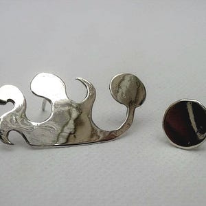 May include: A pair of silver-toned earrings. One earring is a flowing, wave-like design, and the other is a small, round stud with a dark, marbled appearance. The earrings are displayed on a light background.