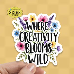 May include: A die-cut sticker featuring the black text "WHERE CREATIVITY BLOOMS WILD" surrounded by a colorful illustration of various wildflowers including pink, yellow, blue, and purple blossoms with green stems and leaves on a white background.