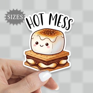 May include: A sticker featuring a cute cartoon s'more with the text "HOT MESS". The s'more has a toasted marshmallow on top of chocolate and graham crackers. The sticker is white with a die-cut shape.