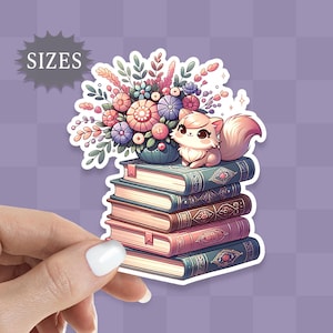 May include: A cute white cat with a pink nose and tail sits on a stack of five colorful books. The cat is surrounded by a bouquet of pink, purple, and blue flowers in a green pot.
