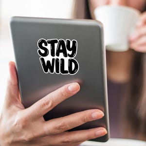 May include: A person is holding a tablet with a black and white sticker that says "Stay Wild".