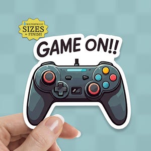 May include: A sticker featuring a cartoon-style video game controller in shades of gray, black, and blue, with red and orange buttons. The text "GAME ON!!" is above the controller. A yellow tag indicates "WATERPROOF SIZES + FINISH".