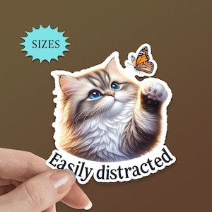 May include: A white and gray fluffy kitten with blue eyes reaches up towards a butterfly with orange and black wings. The text "Easily distracted" is below the kitten.