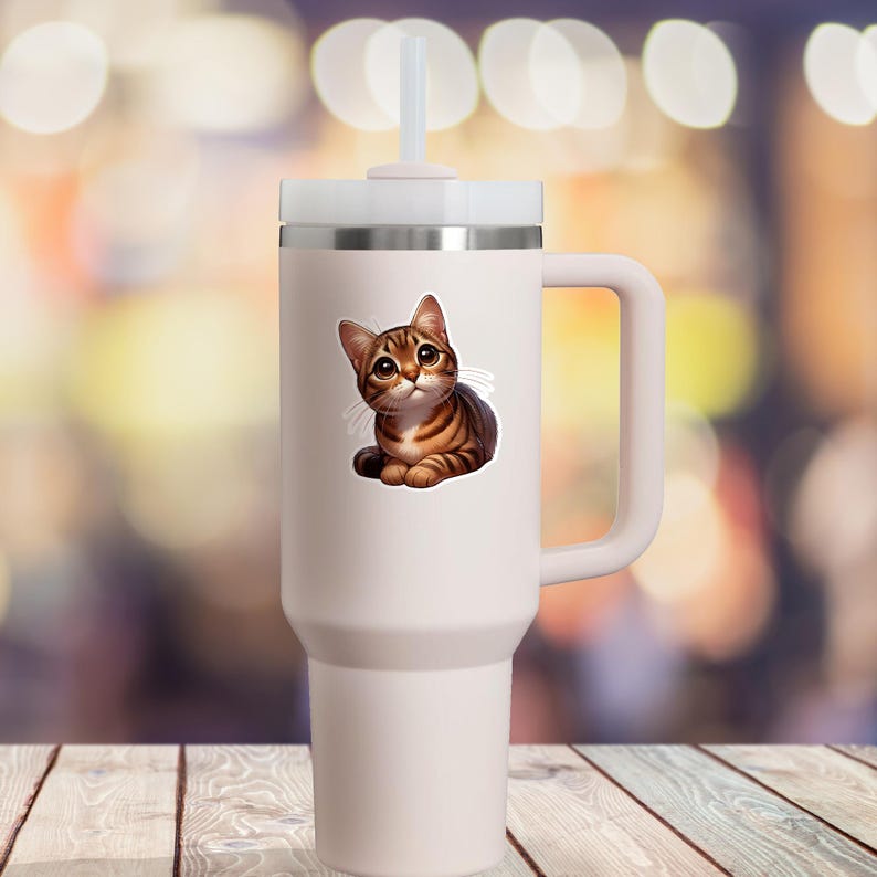 May include: A white travel mug with a brown tabby cat sticker. The cat is looking up with big eyes and has brown and white stripes.