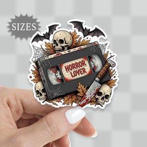 May include: A sticker featuring a black VHS tape with the words "HORROR LOVER" on the label. The design includes skulls, bats, a bloody knife, and autumn leaves, creating a spooky, Halloween theme.