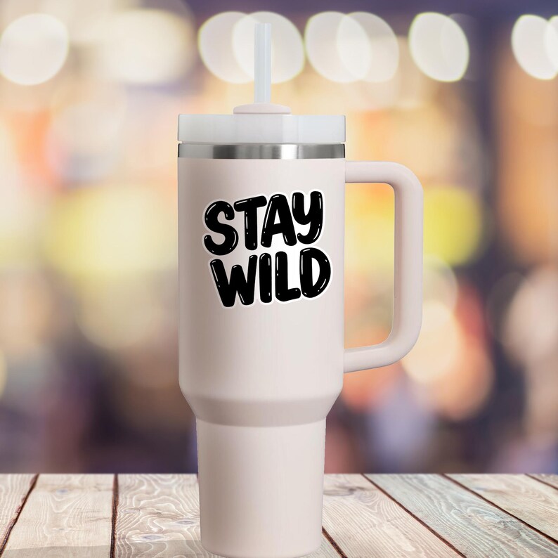 May include: A white travel mug with a black handle and a black lid. The mug has the text "Stay Wild" printed on the front in a black, bubble-like font.