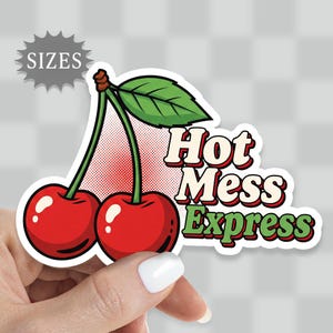 May include: A sticker featuring two bright red cherries with a green leaf and the words "Hot Mess Express" in a retro font. The sticker is held by a hand with a white painted nail.