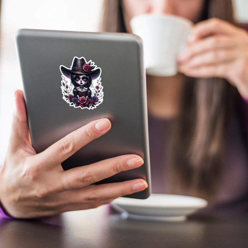 May include: A sticker featuring a digital illustration of a girl with dark hair wearing a black cowboy hat and a black dress. She is surrounded by red and purple flowers.