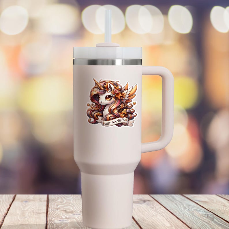 K&ouml;nnte beinhalten: Ein hellrosa isolierter Becher mit wei&szlig;em Deckel und Griff. Auf der Vorderseite befindet sich ein Aufkleber mit einem Einhorn in Herbstfarben und dem Text "AUTUMN VIBES". Der Becher hat einen Strohhalm und ist f&uuml;r hei&szlig;e oder kalte Getr&auml;nke konzipiert.